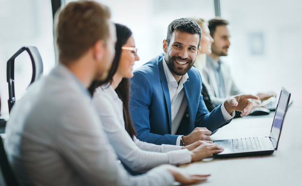 Smiling man in blue suit jacket sitting at a table looking a fellow work mates